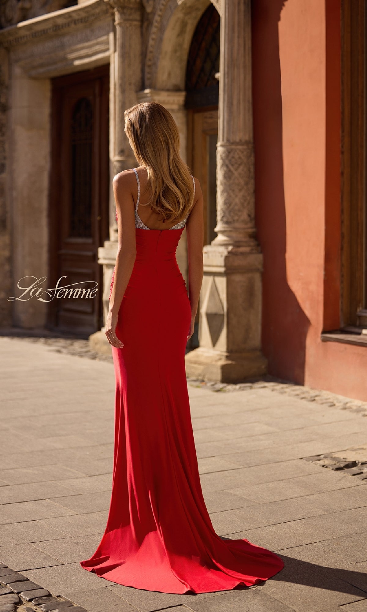 A woman in the Beaded-Neckline Long Prom Dress: La Femme 32939 stands outdoors with her back to the camera in front of an ornate building.